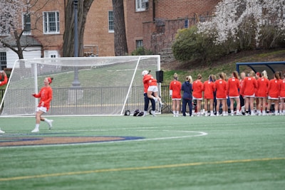 A group of female athletes in orange uniforms stand near a soccer goal on a grassy field. Most of them face away from the viewer, with one player engaging in conversation with a teammate. Trees and a brick building are visible in the background, contributing to a campus-like atmosphere.