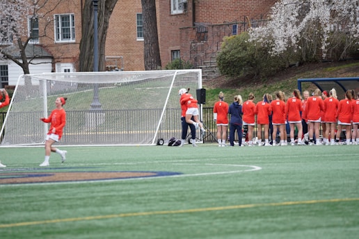 A group of female athletes in orange uniforms stand near a soccer goal on a grassy field. Most of them face away from the viewer, with one player engaging in conversation with a teammate. Trees and a brick building are visible in the background, contributing to a campus-like atmosphere.