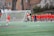 A group of female athletes in orange uniforms stand near a soccer goal on a grassy field. Most of them face away from the viewer, with one player engaging in conversation with a teammate. Trees and a brick building are visible in the background, contributing to a campus-like atmosphere.