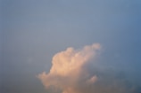 A single fluffy cloud with a soft, warm glow against a clear blue sky. The cloud appears to be illuminated by sunlight, creating a gentle contrast between the warm and cool tones.