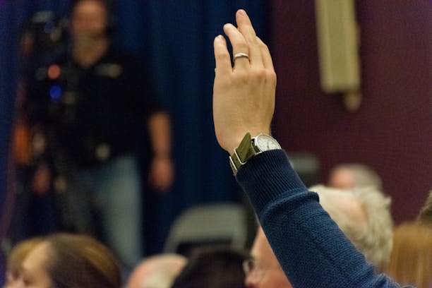 A raised hand wearing a wristwatch is in the foreground, signifying participation or attention in a conference or meeting. The background features blurred figures and a camera setup, with a dark blue curtain and various heads indicating an audience setting.
