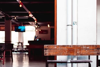 The interior features a modern, industrial design with exposed pipes and hanging light bulbs. A wooden bench with 'VERSATILE MINDS' etched on it is prominently placed, and there is a dimly lit bar area in the background. A television is mounted on the wall, and several empty bar stools are scattered throughout the space.