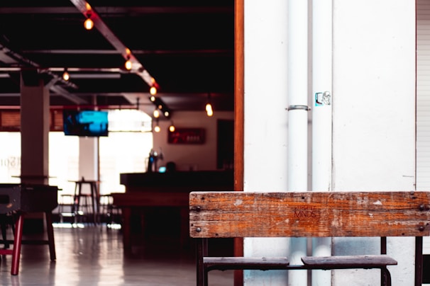 The interior features a modern, industrial design with exposed pipes and hanging light bulbs. A wooden bench with 'VERSATILE MINDS' etched on it is prominently placed, and there is a dimly lit bar area in the background. A television is mounted on the wall, and several empty bar stools are scattered throughout the space.