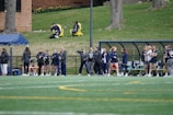 A group of female athletes, some in sports uniforms and others in jackets, are gathered along the sideline of a sports field. They seem to be in a team environment, possibly preparing for or observing a game. In the background, there are a few people sitting on yellow chairs on a grassy incline, and a building with a brick facade.