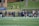 A group of female athletes, some in sports uniforms and others in jackets, are gathered along the sideline of a sports field. They seem to be in a team environment, possibly preparing for or observing a game. In the background, there are a few people sitting on yellow chairs on a grassy incline, and a building with a brick facade.