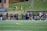 A group of female athletes, some in sports uniforms and others in jackets, are gathered along the sideline of a sports field. They seem to be in a team environment, possibly preparing for or observing a game. In the background, there are a few people sitting on yellow chairs on a grassy incline, and a building with a brick facade.