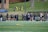 A group of female athletes, some in sports uniforms and others in jackets, are gathered along the sideline of a sports field. They seem to be in a team environment, possibly preparing for or observing a game. In the background, there are a few people sitting on yellow chairs on a grassy incline, and a building with a brick facade.