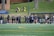 A group of female athletes, some in sports uniforms and others in jackets, are gathered along the sideline of a sports field. They seem to be in a team environment, possibly preparing for or observing a game. In the background, there are a few people sitting on yellow chairs on a grassy incline, and a building with a brick facade.