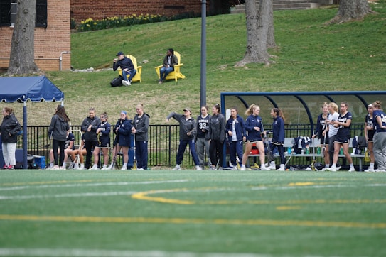 A group of female athletes, some in sports uniforms and others in jackets, are gathered along the sideline of a sports field. They seem to be in a team environment, possibly preparing for or observing a game. In the background, there are a few people sitting on yellow chairs on a grassy incline, and a building with a brick facade.