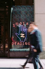 A neon sign with a large hand symbol is displayed in a shop window, featuring bold letters spelling out 'ZODIAC READING.' The background is decorated with a colorful pattern, and two people are walking by, blurred in motion, adding a sense of urban energy.