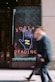 A neon sign with a large hand symbol is displayed in a shop window, featuring bold letters spelling out 'ZODIAC READING.' The background is decorated with a colorful pattern, and two people are walking by, blurred in motion, adding a sense of urban energy.