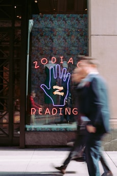 A neon sign with a large hand symbol is displayed in a shop window, featuring bold letters spelling out 'ZODIAC READING.' The background is decorated with a colorful pattern, and two people are walking by, blurred in motion, adding a sense of urban energy.