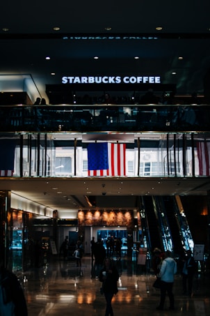 Inside a shopping mall, there are people walking under a Starbucks Coffee shop on the upper level. An American flag is prominently displayed, and the floor is shiny and reflective with warm lighting. The scene includes escalators and a variety of retail elements.