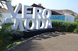 Large white letters spelling 'VERO MODA' are displayed on a black platform in an outdoor setting. Surrounding the sign are green bushes and palm-like plants. A blue building with the word 'Soleil' is in the background, along with brown hills and a clear sky.
