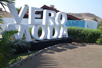 Large white letters spelling 'VERO MODA' are displayed on a black platform in an outdoor setting. Surrounding the sign are green bushes and palm-like plants. A blue building with the word 'Soleil' is in the background, along with brown hills and a clear sky.