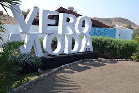 Large white letters spelling 'VERO MODA' are displayed on a black platform in an outdoor setting. Surrounding the sign are green bushes and palm-like plants. A blue building with the word 'Soleil' is in the background, along with brown hills and a clear sky.