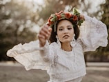 A person wearing a white lace dress adorned with floral earrings and a vibrant red flower crown, set against a blurred natural background.