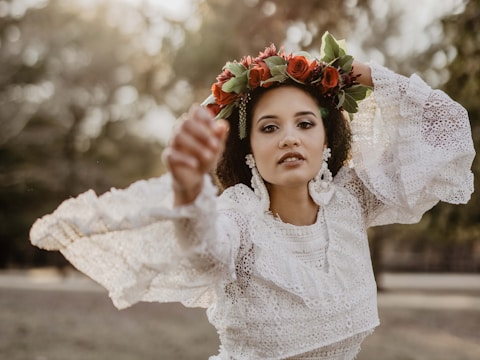 A person wearing a white lace dress adorned with floral earrings and a vibrant red flower crown, set against a blurred natural background.