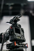 A professional camera setup with a mounted digital camera on a tripod. Various attachments and cables are visible, including a lens with a wide aperture, an antenna, and a control panel with buttons and dials.