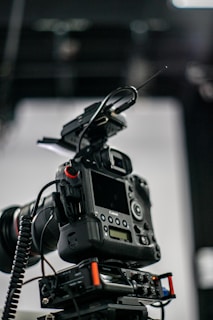 A professional camera setup with a mounted digital camera on a tripod. Various attachments and cables are visible, including a lens with a wide aperture, an antenna, and a control panel with buttons and dials.
