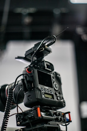 A professional camera setup with a mounted digital camera on a tripod. Various attachments and cables are visible, including a lens with a wide aperture, an antenna, and a control panel with buttons and dials.