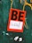 A vibrant red sticker with black letters spelling the word 'BE' is placed on a textured teal surface. Below it, a white section contains the word 'LOVE' with an arrow pointing upward. Yellow abstract paint markings are visible in the background.