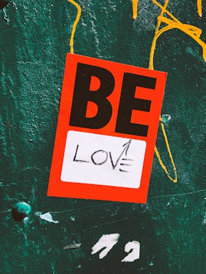 A vibrant red sticker with black letters spelling the word 'BE' is placed on a textured teal surface. Below it, a white section contains the word 'LOVE' with an arrow pointing upward. Yellow abstract paint markings are visible in the background.