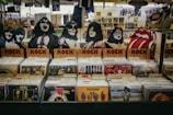 A vinyl record store display featuring cardboard cutouts of band members and symbols, with labeled sections for rock albums. Various album covers, including recognizable bands, are visible. The background shows more records and music memorabilia.