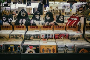 A vinyl record store display featuring cardboard cutouts of band members and symbols, with labeled sections for rock albums. Various album covers, including recognizable bands, are visible. The background shows more records and music memorabilia.