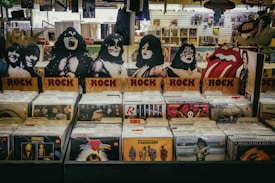 A vinyl record store display featuring cardboard cutouts of band members and symbols, with labeled sections for rock albums. Various album covers, including recognizable bands, are visible. The background shows more records and music memorabilia.
