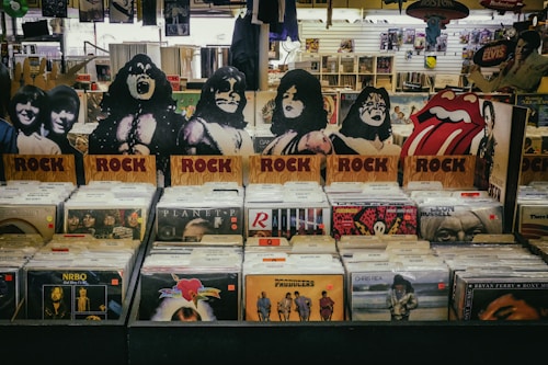 A vinyl record store display featuring cardboard cutouts of band members and symbols, with labeled sections for rock albums. Various album covers, including recognizable bands, are visible. The background shows more records and music memorabilia.
