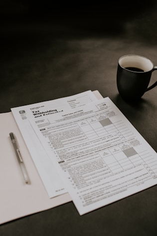 A set of tax forms and documents is placed neatly on a dark surface. There is a black coffee mug to the right of the documents, and a pen is resting on a folder. The scene suggests a workspace setting.