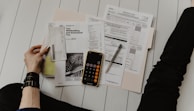 A person is sitting on the floor surrounded by tax forms and documents, holding a piece of paper in one hand. There is a smartphone with a calculator app open, and a pen placed on one of the documents.