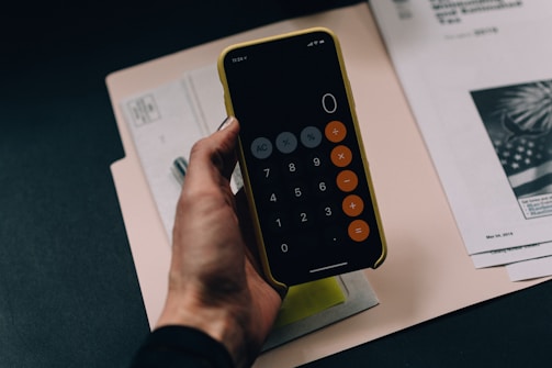 A close-up of a hand holding a smartphone with a calculator app open, showing the number zero. Papers and documents are scattered underneath on a dark table, including a visible American flag on one of the documents.