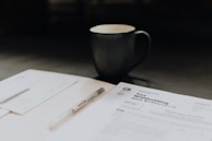 The scene includes a dimly lit workspace with a focus on tax-related documents, a pen, and a mug. The documents are prominently featured on the table, indicating a financial or tax preparation setting.