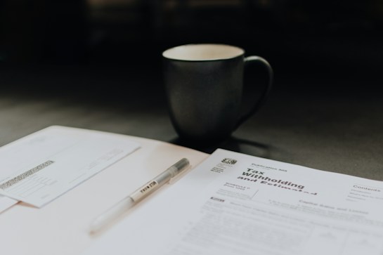 The scene includes a dimly lit workspace with a focus on tax-related documents, a pen, and a mug. The documents are prominently featured on the table, indicating a financial or tax preparation setting.