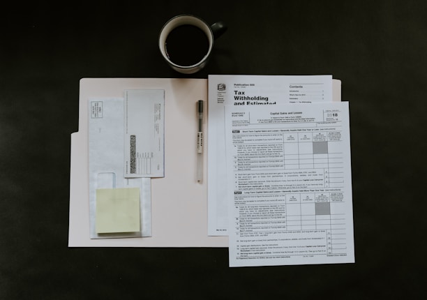 A neatly arranged setup featuring tax documents on top of a light-colored folder. An envelope and a sticky note are placed on the left side. A black pen is positioned near the paperwork. A coffee cup sits above the documents, suggesting a moment of work or review.