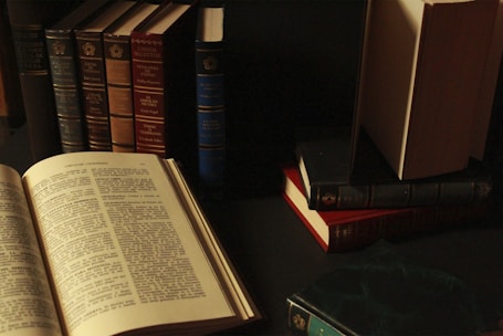 A collection of vintage hardcover books arranged on a dark surface, with some standing vertically and others stacked horizontally. An open book is prominently displayed in the foreground, revealing text on the pages. The lighting is dim, creating a cozy and scholarly atmosphere.