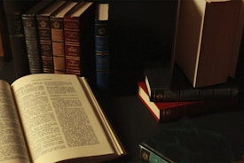 A collection of vintage hardcover books arranged on a dark surface, with some standing vertically and others stacked horizontally. An open book is prominently displayed in the foreground, revealing text on the pages. The lighting is dim, creating a cozy and scholarly atmosphere.