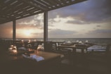 A serene seaside restaurant setting with neatly arranged tables and chairs under a pergola. The sunset casts a warm glow over the ocean as the sky transitions from blue to shades of orange and purple. Each table is set with glasses and napkins, creating a peaceful atmosphere.