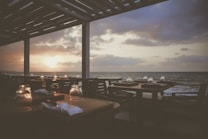 A serene seaside restaurant setting with neatly arranged tables and chairs under a pergola. The sunset casts a warm glow over the ocean as the sky transitions from blue to shades of orange and purple. Each table is set with glasses and napkins, creating a peaceful atmosphere.