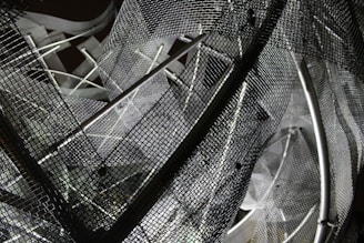 A complex arrangement of metal mesh wires and rods creating an abstract geometric pattern. The mesh creates a layered, intricate design with overlapping grids and shadows, reflecting various shades of gray.