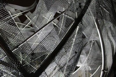 A complex arrangement of metal mesh wires and rods creating an abstract geometric pattern. The mesh creates a layered, intricate design with overlapping grids and shadows, reflecting various shades of gray.
