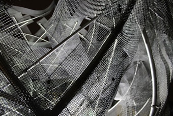 A complex arrangement of metal mesh wires and rods creating an abstract geometric pattern. The mesh creates a layered, intricate design with overlapping grids and shadows, reflecting various shades of gray.