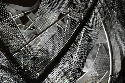 A complex arrangement of metal mesh wires and rods creating an abstract geometric pattern. The mesh creates a layered, intricate design with overlapping grids and shadows, reflecting various shades of gray.