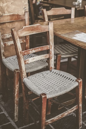 Two rustic wooden chairs with woven seats are placed next to a wooden table. The chairs and table have a worn, vintage look, suggesting an antique or retro setting. The texture of the woven seats contrasts with the weathered wood of the chairs.