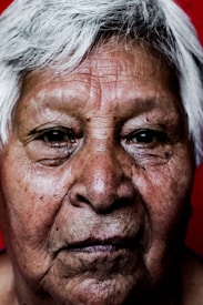 A close-up portrait of an elderly person with white hair, showcasing deeply wrinkled skin and prominent features. The background is a solid red color, providing a stark contrast to the person's complexion.