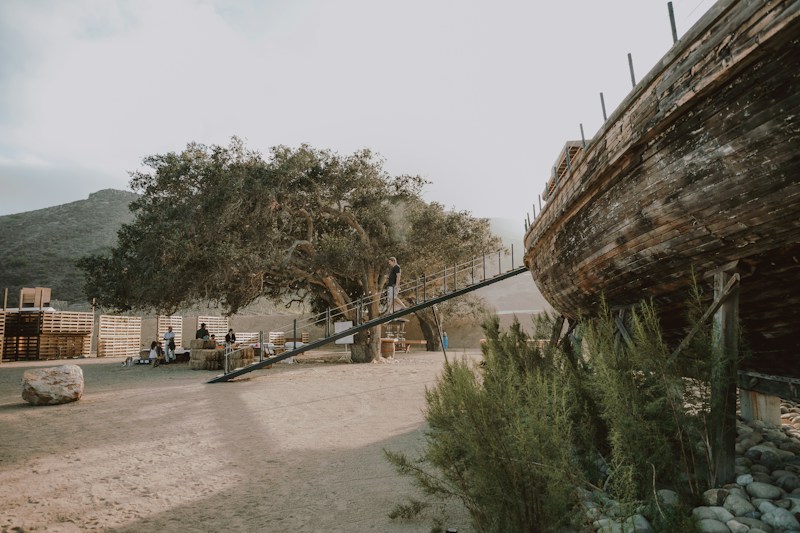A rustic outdoor setting with a large tree in the center, where people are gathered around. On the right side, an old, weathered wooden ship is elevated, with a ramp leading up to it. The background features hills and a partly cloudy sky creating a calm and natural atmosphere. Several wooden pallets are arranged near the tree, giving a casual and rustic vibe.