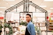 A person stands in a store aisle surrounded by various home decor items. There are small potted plants, rolled towels, and jars on the shelves. The area is lit with bright lights from the ceiling.