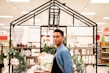 A person stands in a store aisle surrounded by various home decor items. There are small potted plants, rolled towels, and jars on the shelves. The area is lit with bright lights from the ceiling.